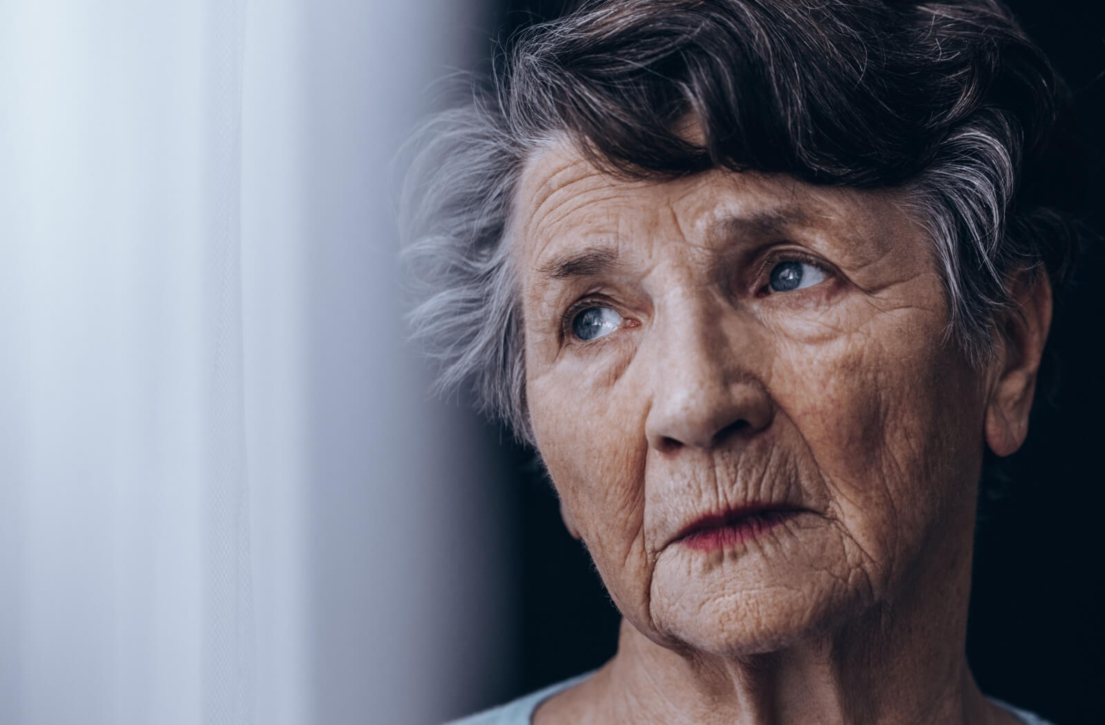 An older adult woman looking out the window with a serious expression.