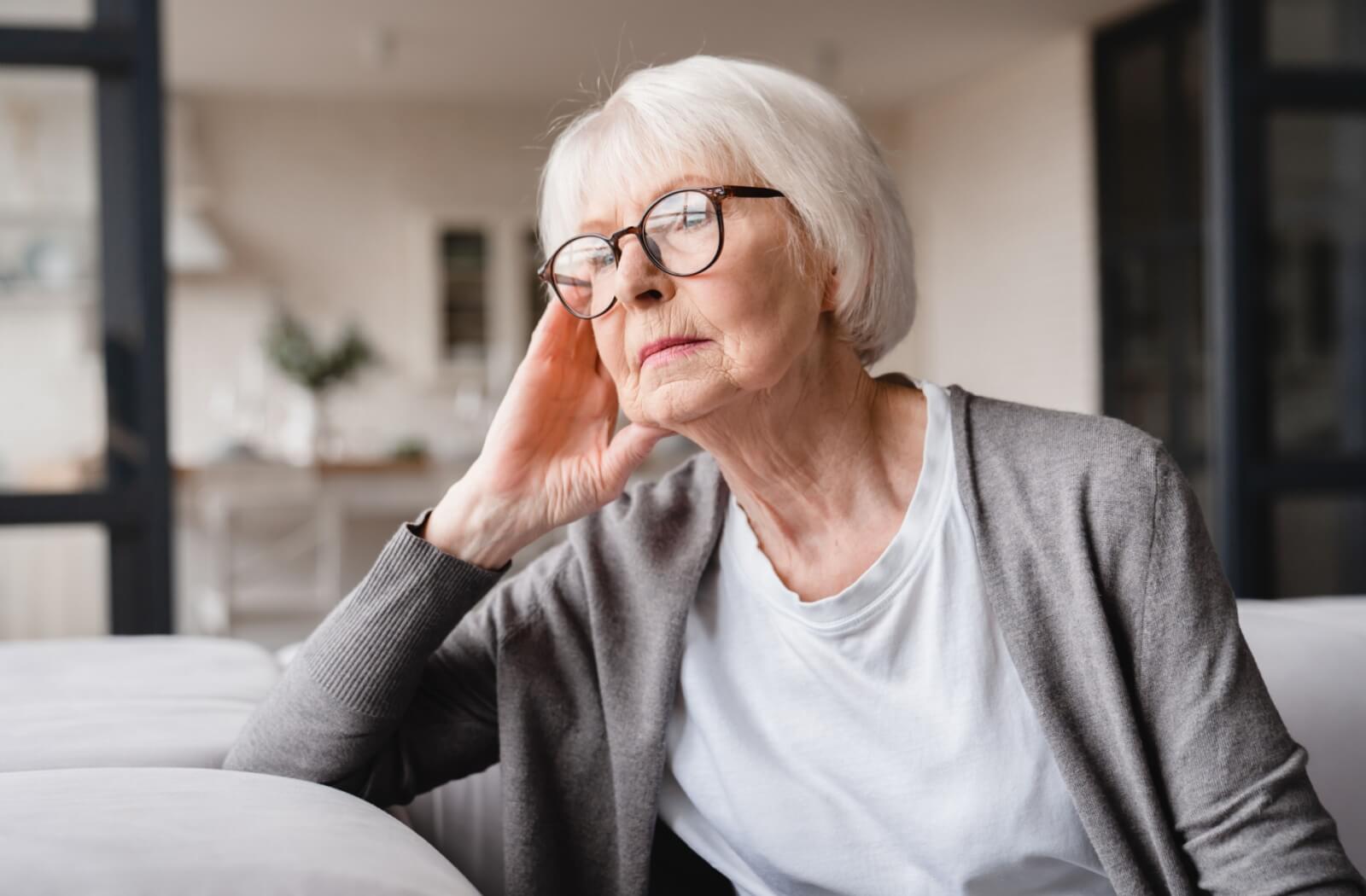 A senior with glasses on sitting on a couch and looking out the window with a serious expression