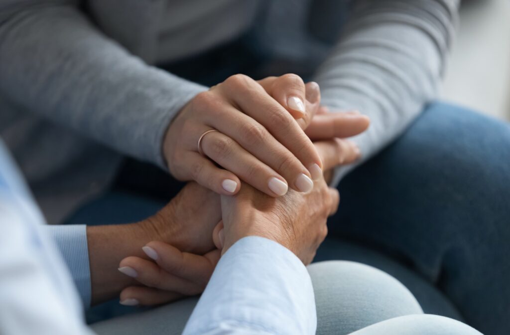 A close-up of a daughter holding her elderly mother’s hands as they sit together on a couch in a senior community.