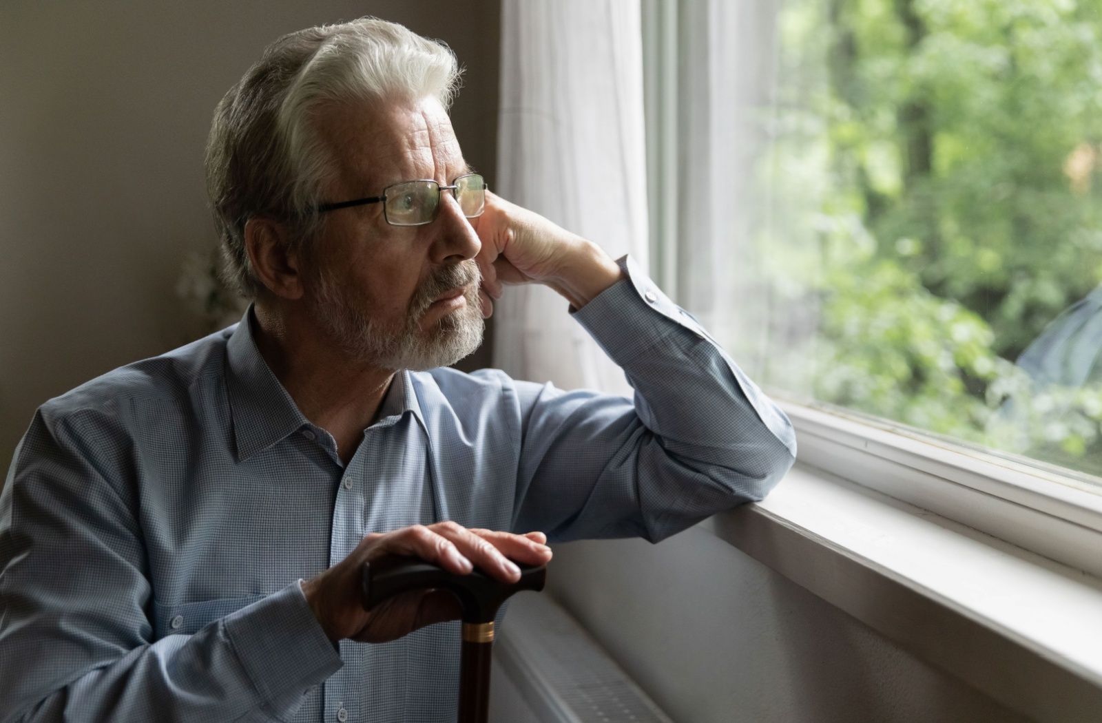 An older adult man with glasses leaning on a windowsill and resting his right hand on the top of a cane while looking out the window with a serious expression.