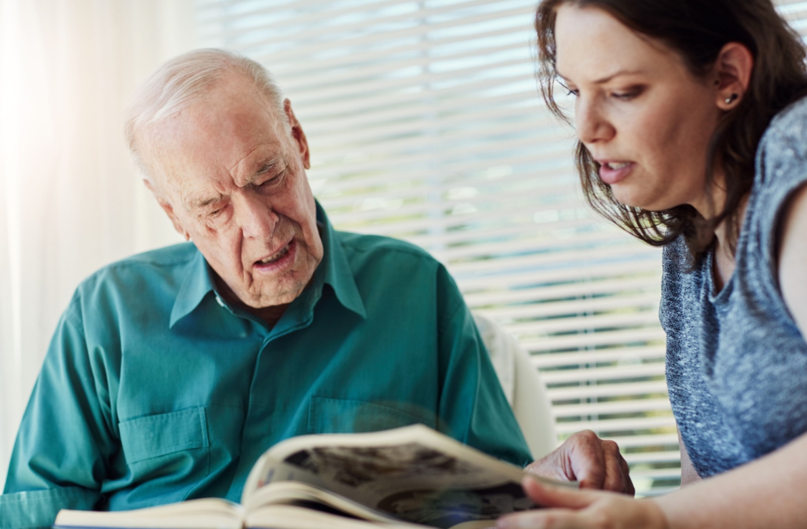 Family member helping an elderly man with Alzheimer's look through a photo album, offering support and engagement through memory recall.