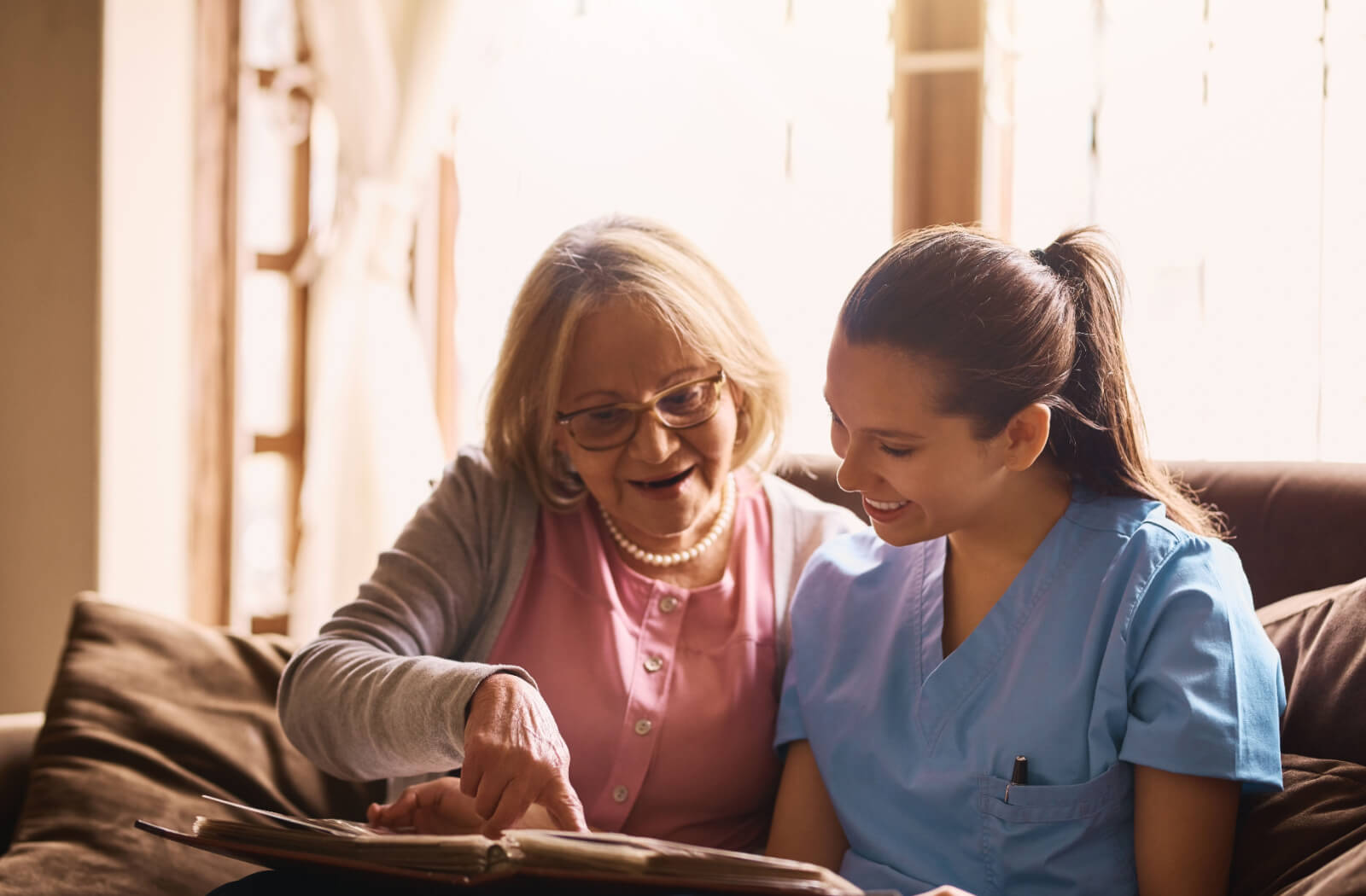 An older adult in memory care showing off pictures in a photo album to a caregiver during a reminiscence therapy exercise.
