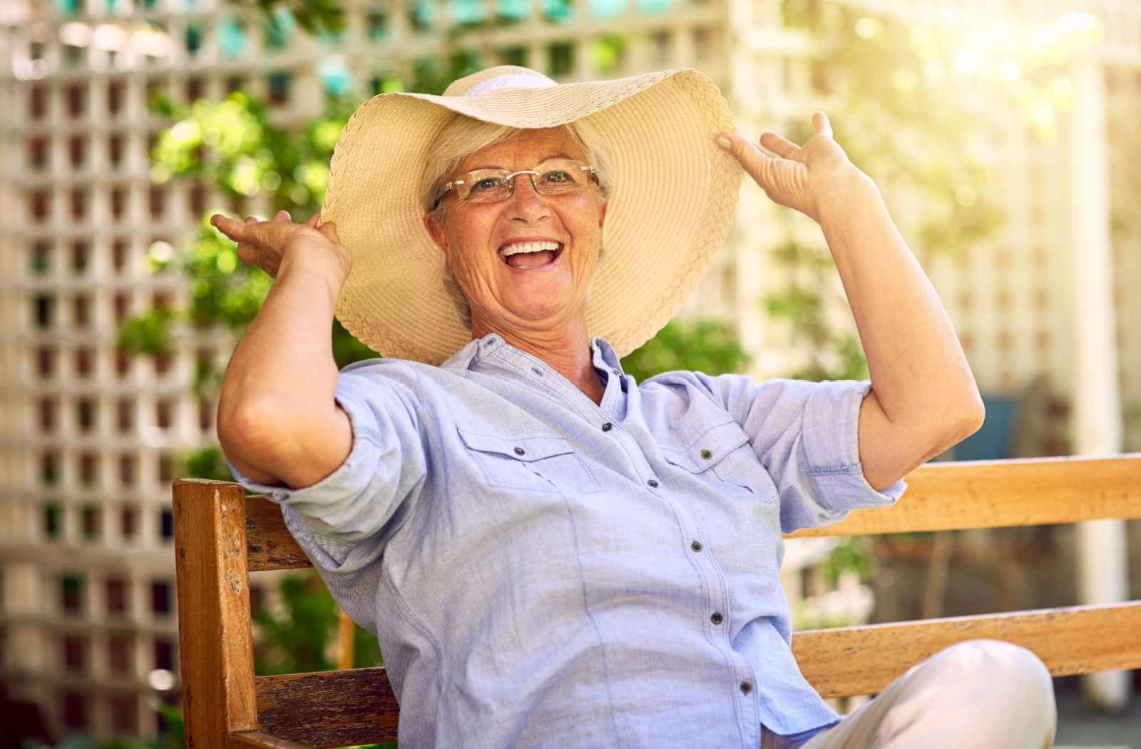 A laughing older adult in a sunhat, sitting on a wooden bench in the gardens of a memory care community.