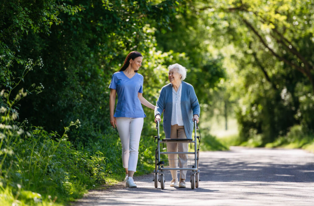 An older adult using a walker, smiling at a caregiver in appreciation during a summertime walk through the community gardens.
