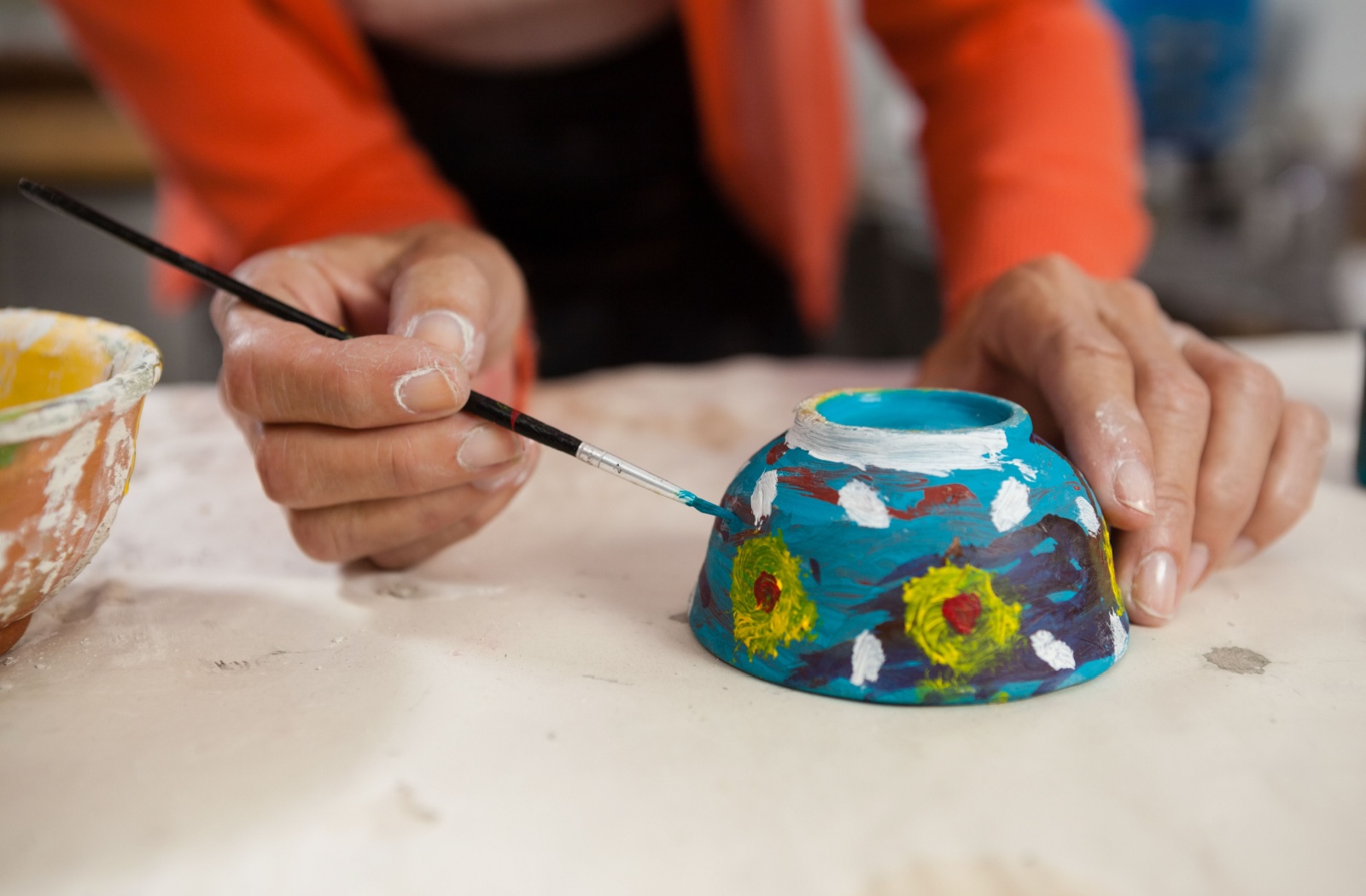 A close-up image of an older adult's hands as they paint small flowers on a blue bowl.