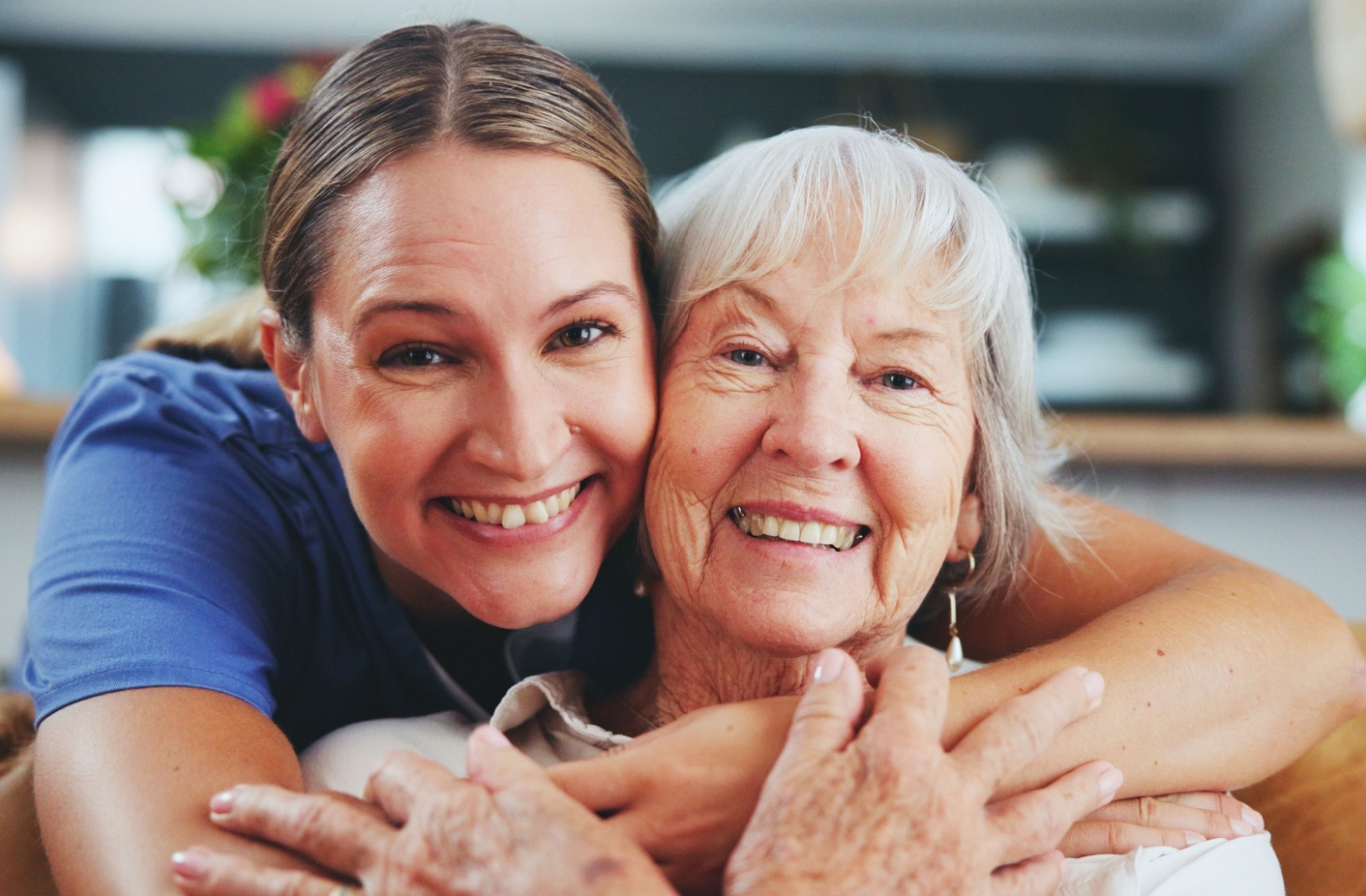 A caregiver laughs with a senior dressed in pink and pearls inside a comfortable unit in a memory care community.