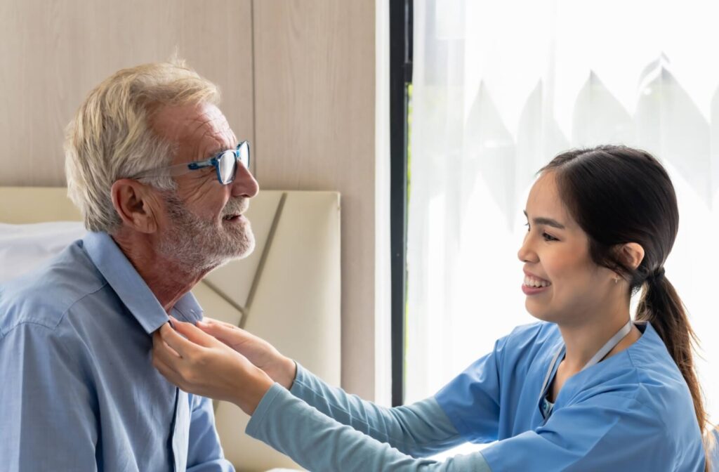 A smiling young caregiver, dressed in blue scrubs, assists a senior with adjusting their shirt collar in a bright memory care setting.