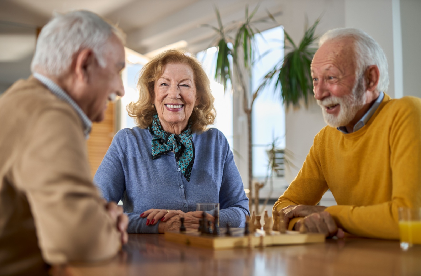 A group of older adults laugh together during a chess game in a common lounge of a warm senior living community