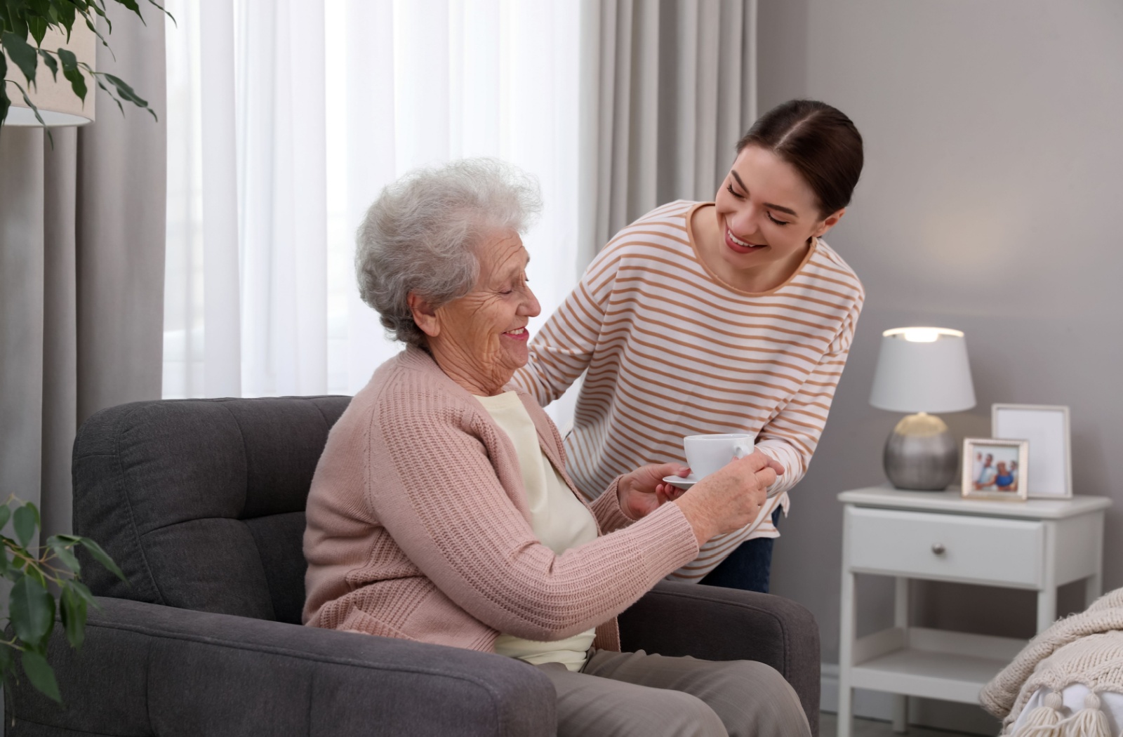 A young caregiver brings an older adult a mug of tea in their living room in memory care to help them maintain their daily routine